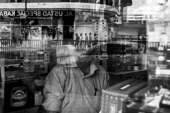 Black-and-white double exposure of a shopkeeper and a busy street